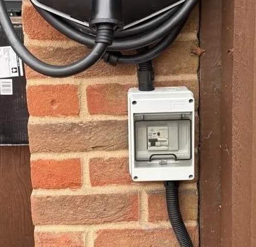 A charger’s protection box and cable run on a brick pier beside the garage