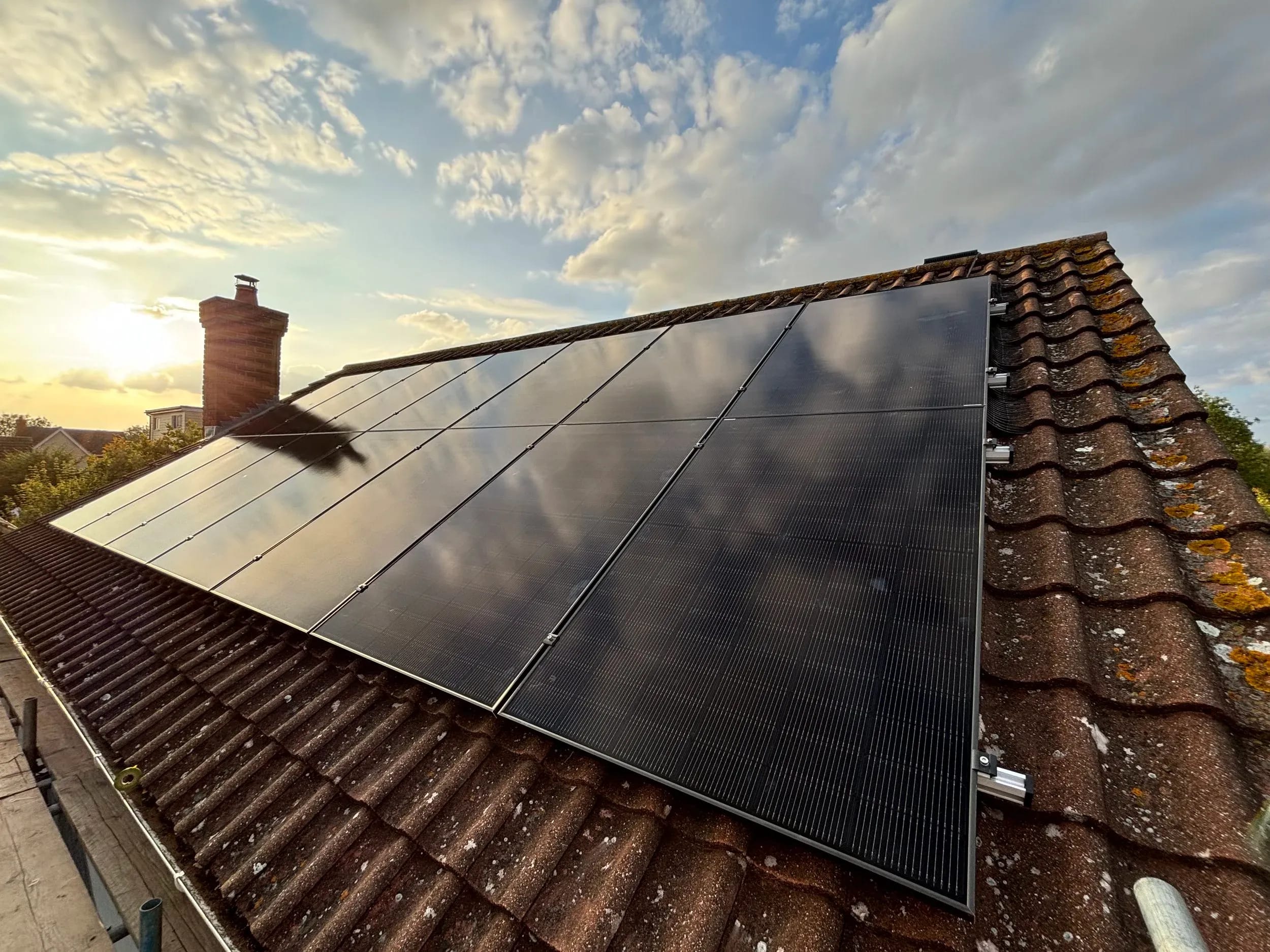 Fourteen black panels on a tiled roof in low evening sun, a chimney beside them