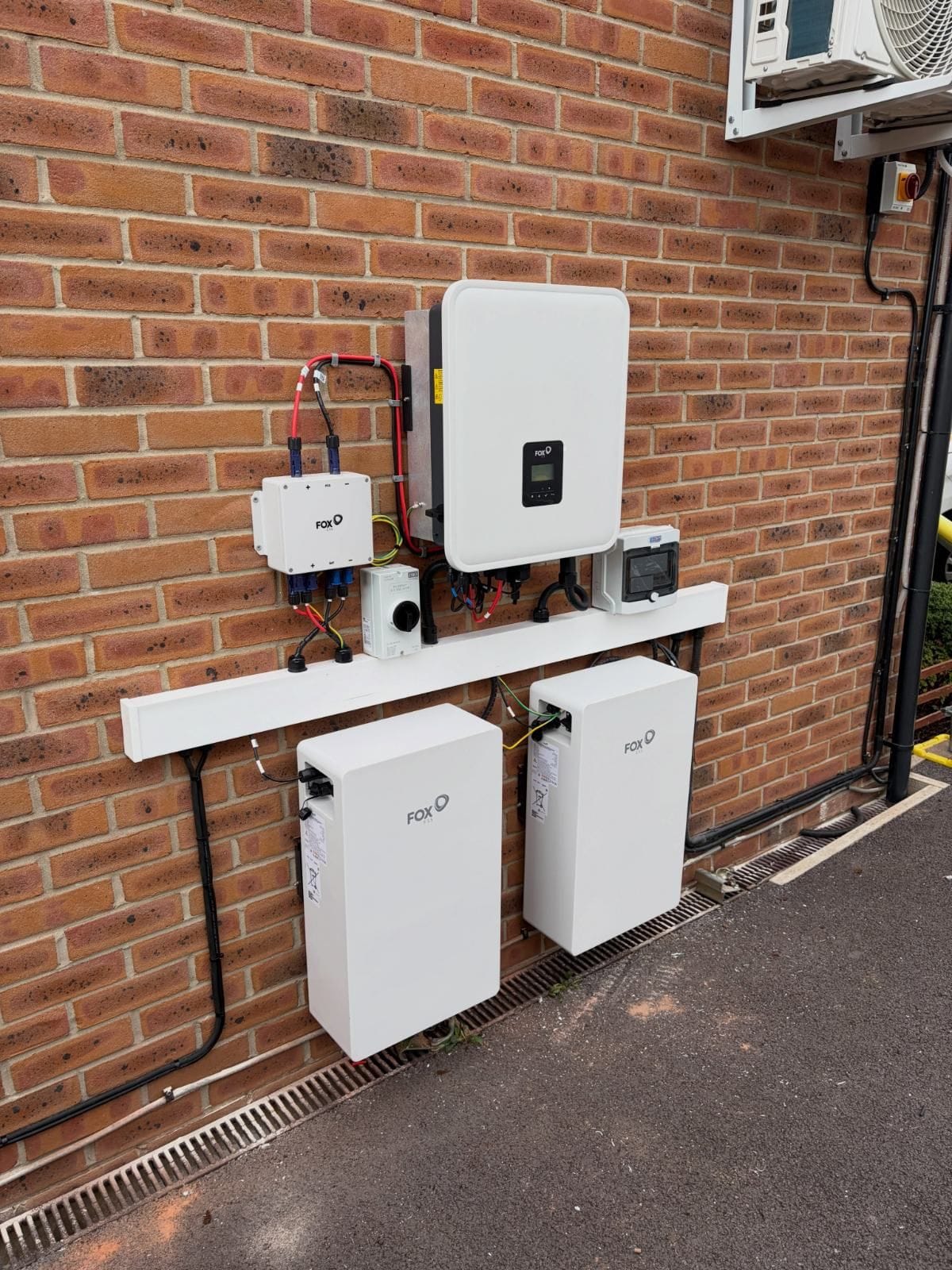 An inverter and two battery units fitted to the brick wall of a house, isolators on white trunking between them